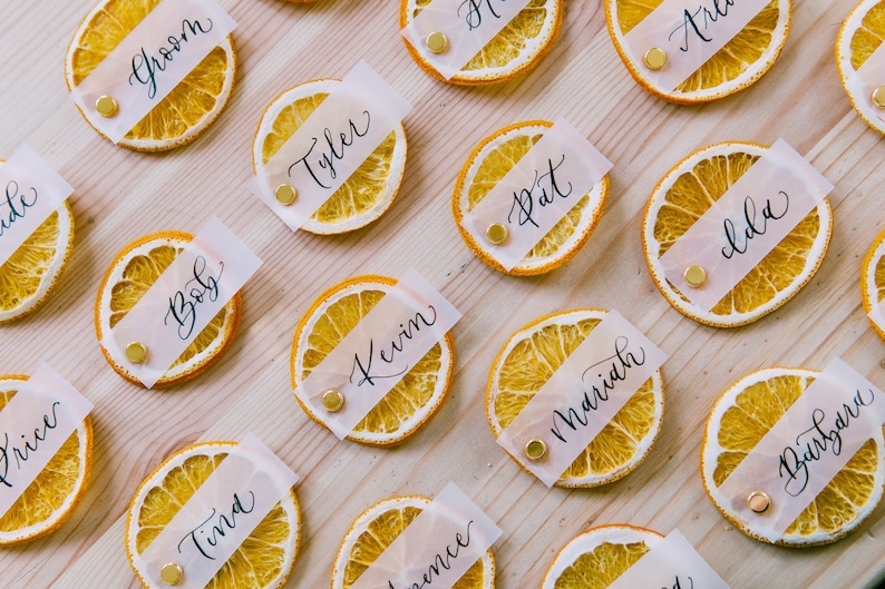 May include: Dried orange slices with white paper place cards with names written in black calligraphy. The names include Groom, Tyler, Pat, Ida, Bob, Kevin, Mariah, Barbara, Tina, and Price.