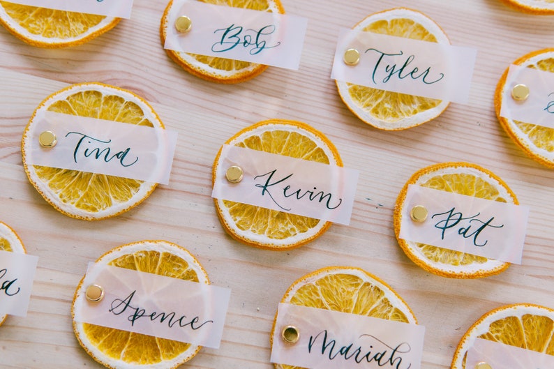 May include: Dried orange slices with white paper place cards with names written in black calligraphy. The names are Tina, Bob, Tyler, Kevin, Pat, Spence, and Mariah.