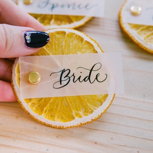 May include: Dried orange slice place card with a gold push pin and a white card with the word "Bride" written in black calligraphy.
