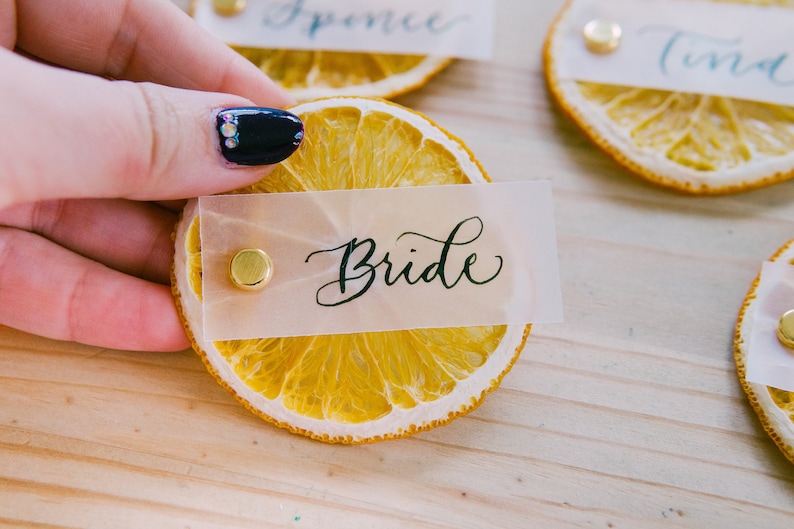 May include: Dried orange slice place card with a gold push pin and a white card with the word "Bride" written in black calligraphy.