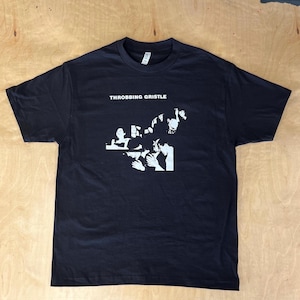 Op de afbeelding: Zwart T-shirt met een witte grafische print van een band genaamd "Throbbing Gristle".