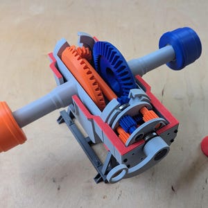 Op de afbeelding: Een doorsnedemodel van een tandwielsysteem met oranje en blauwe tandwielen. Het model is gemaakt van grijs plastic en heeft een rode basis. De tandwielen zijn zichtbaar door de doorsnede van het model.
