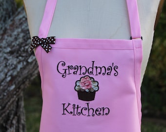 Puede incluir: Delantal rosa con las palabras "Grandma's Kitchen" bordadas y un diseño de cupcake. El delantal tiene un lazo de lunares marrón y blanco y una correa ajustable para el cuello. Ideal para cocinar.