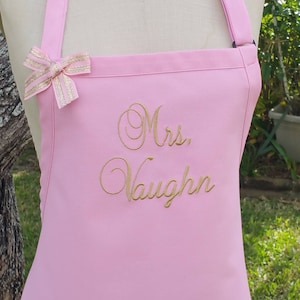 May include: A light pink apron with the embroidered text "Mrs. Vaughn" in gold script. The apron has a bow with gold stripes and adjustable straps. The apron is designed for kitchen or craft use.