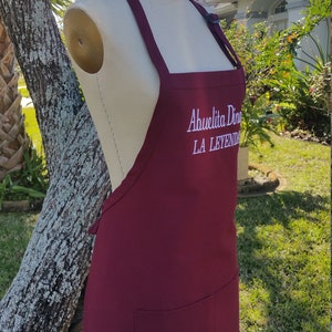 Puede incluir: Delantal burdeos con un bolsillo y el texto bordado "Abuelita Dina LA LEYENDA" en blanco. El delantal se exhibe en un maniqu&iacute; al aire libre, con un &aacute;rbol y vegetaci&oacute;n en el fondo.