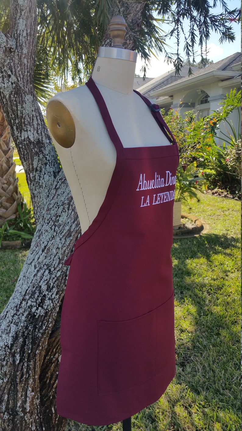 Puede incluir: Delantal burdeos con un bolsillo y el texto bordado "Abuelita Dina LA LEYENDA" en blanco. El delantal se exhibe en un maniqu&iacute; al aire libre, con un &aacute;rbol y vegetaci&oacute;n en el fondo.