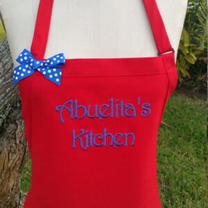 Puede incluir: Un delantal rojo con el texto bordado "Abuelita's Kitchen" en azul. Un lazo azul con lunares blancos est&aacute; sujeto a la correa del delantal. El delantal se muestra en un maniqu&iacute;.