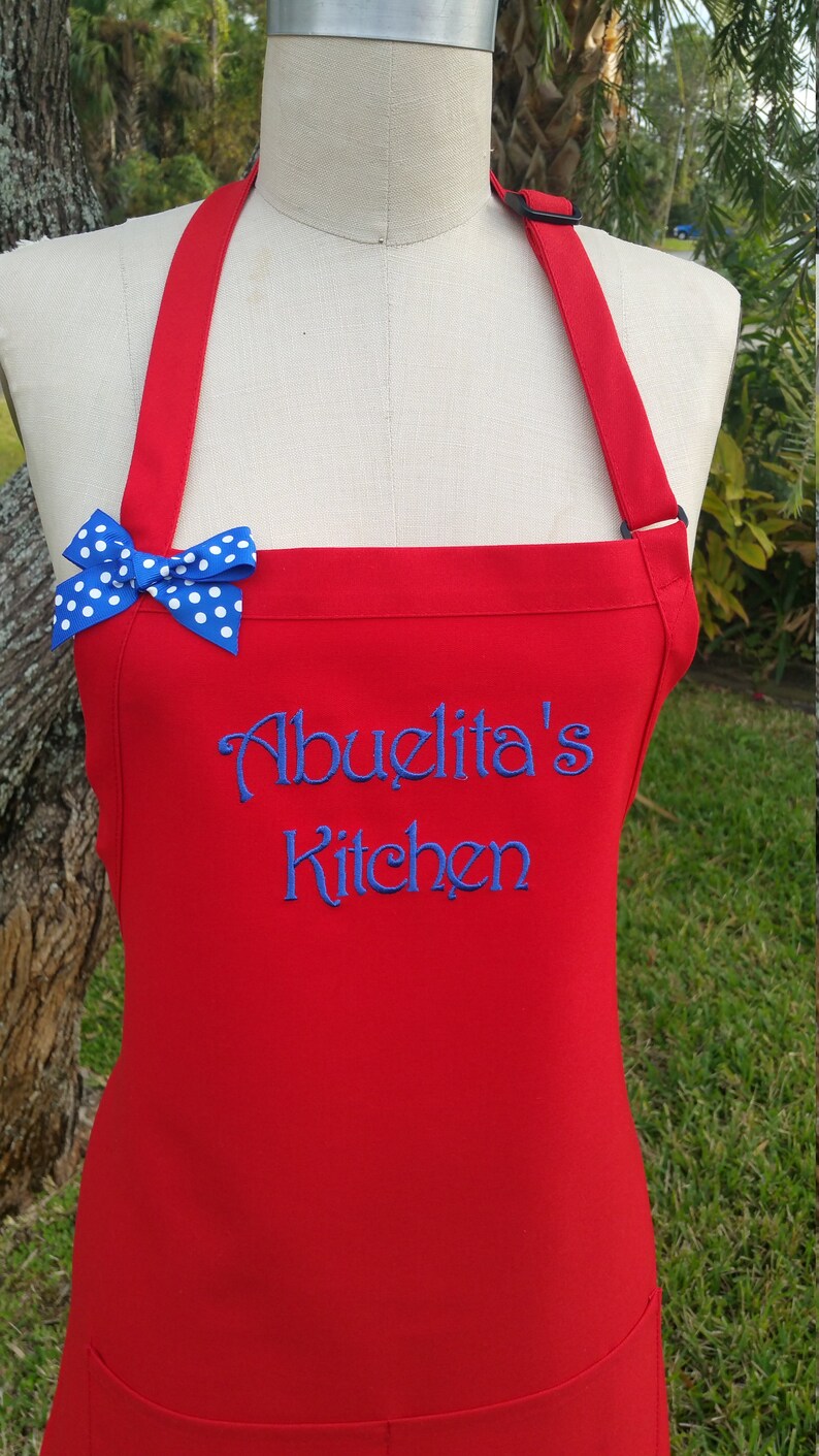 Puede incluir: Un delantal rojo con el texto bordado "Abuelita's Kitchen" en azul. Un lazo azul con lunares blancos est&aacute; sujeto a la correa del delantal. El delantal se muestra en un maniqu&iacute;.