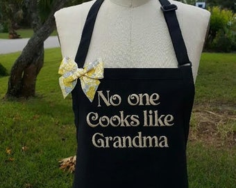 Puede incluir: Delantal negro con el texto bordado "No one cooks like Grandma". Un lazo con estampado amarillo y blanco está prendido. El delantal se exhibe en un maniquí.