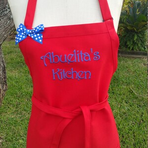 Puede incluir: Delantal rojo con la inscripci&oacute;n bordada en azul "Abuelita's Kitchen". Un lazo azul con lunares blancos adorna el peto. El delantal tiene una cinta en la cintura y dos bolsillos delanteros.