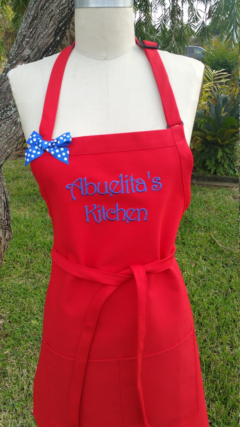 Puede incluir: Delantal rojo con la inscripci&oacute;n bordada en azul "Abuelita's Kitchen". Un lazo azul con lunares blancos adorna el peto. El delantal tiene una cinta en la cintura y dos bolsillos delanteros.