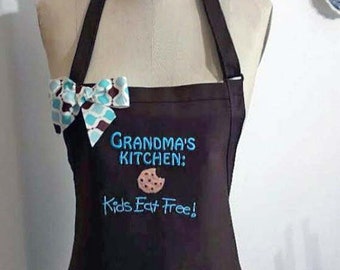 Puede incluir: Un delantal marrón oscuro con un lazo y el texto "GRANDMA'S KITCHEN: Kids Eat Free!" bordado en azul. Un gráfico de galleta también está bordado en el delantal. El lazo presenta un patrón blanco, azul y granate.