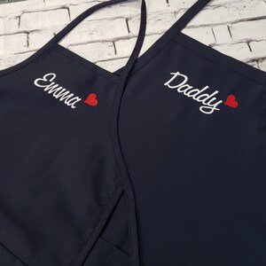 Puede incluir: Dos delantales azul marino con bordados blancos y un corazón rojo. Uno dice "Emma" y el otro "Daddy". Los delantales tienen correas largas para atar alrededor del cuello y la cintura.