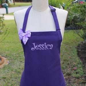 May include: A purple apron with the name "Jessica" embroidered in cursive. The apron has a bow with white polka dots. The apron is displayed on a mannequin outdoors.