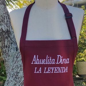 Puede incluir: Un delantal burdeos con texto bordado en blanco que dice "Abuelita Dina LA LEYENDA". El delantal tiene una correa para el cuello con una hebilla negra ajustable. El delantal se muestra en un maniqu&iacute;.