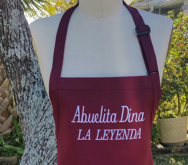 Puede incluir: Un delantal burdeos con texto bordado en blanco que dice "Abuelita Dina LA LEYENDA". El delantal tiene una correa para el cuello con una hebilla negra ajustable. El delantal se muestra en un maniqu&iacute;.