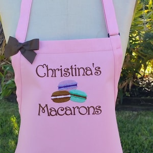 May include: A pink apron with the embroidered text "Christina's Macarons" and three colorful macarons. A brown bow is attached to the apron strap. The apron is designed for kitchen use.