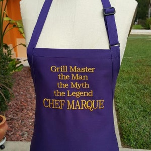 May include: A purple apron with the embroidered text "Grill Master the Man the Myth the Legend CHEF MARQUE". The apron has adjustable straps and a black buckle. The apron is displayed on a mannequin.