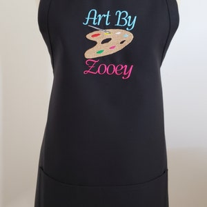 May include: Black apron with the text "Art By Zooey" embroidered in teal and pink. A brown and tan artist's palette with colorful paint is embroidered above the name. The apron has a pocket and adjustable neck strap.