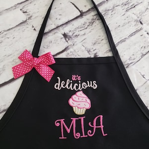 May include: Black apron with a pink polka dot bow and straps. The apron features the embroidered text "it's delicious" above a pink cupcake design. Below the cupcake is the name "MIA" embroidered in pink.