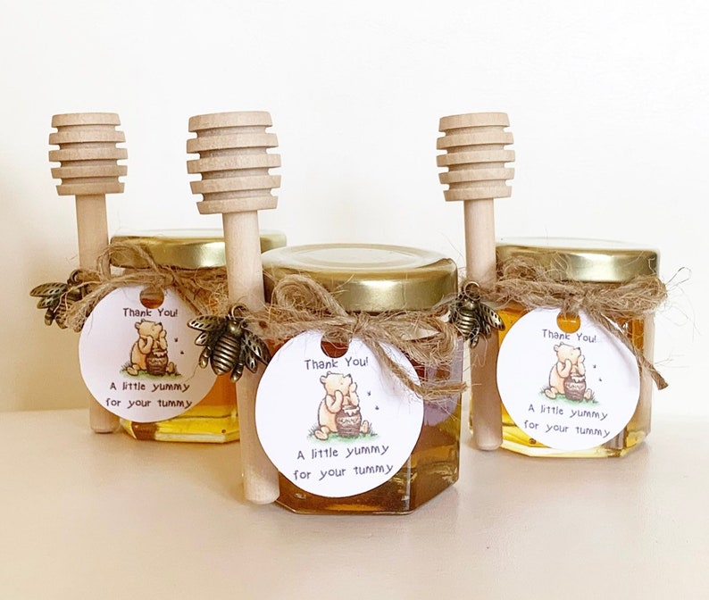 Pode incluir: Tr&ecirc;s pequenos frascos de vidro cheios de mel, cada um com um palito de mel de madeira e uma etiqueta redonda anexada. As etiquetas apresentam um personagem de desenho animado do Winnie the Pooh e o texto "Obrigado! Um pouco delicioso para sua barriga".