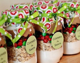 May include: Glass jars filled with cookie ingredients, including oats, chocolate chips, and colorful candies. Each jar is sealed with a festive lid and tied with a green polka-dot ribbon. The label reads "Cookies For Santa."