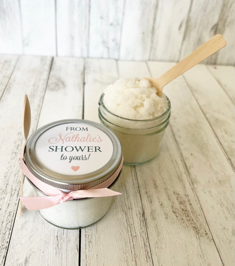 May include: Two small glass jars filled with white sugar scrub, tied with pink ribbon and a small wooden spoon. The label on the jar reads "From Nathalie's Shower to yours!"
