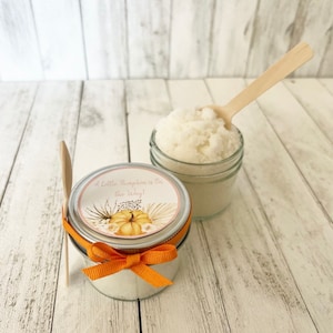 May include: Two glass jars filled with white sugar scrub, one with a label that reads "A Little Pumpkin is on the Way!" with a watercolour pumpkin and floral design. Both jars have wooden spoons.