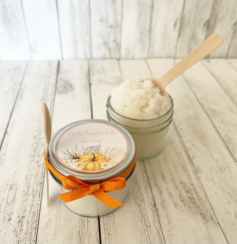 May include: Two glass jars filled with white sugar scrub, one with a label that reads "A Little Pumpkin is on the Way!" with a watercolour pumpkin and floral design. Both jars have wooden spoons.