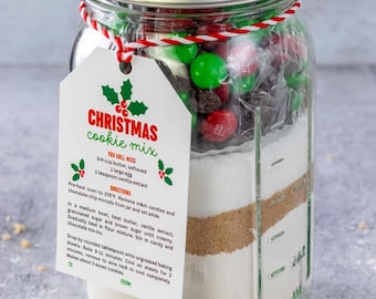 May include: A clear glass mason jar filled with layered dry ingredients for Christmas cookies, topped with red and green candies. A white tag tied with red and white twine displays the text Christmas cookie mix, ingredients, and baking directions.