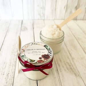 May include: Two small glass jars filled with white sugar scrub, one with a floral patterned label that reads "Chloe & Michael thank you for sharing our day 11.09.99" and a red ribbon tied around the lid, and the other with a wooden spoon inside.
