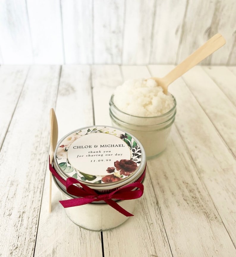 May include: Two small glass jars filled with white sugar scrub, one with a floral patterned label that reads "Chloe & Michael thank you for sharing our day 11.09.99" and a red ribbon tied around the lid, and the other with a wooden spoon inside.