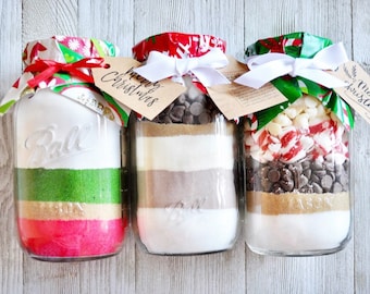 May include: Three glass jars filled with layered ingredients for baking cookies. Each jar is topped with a festive fabric lid, a white ribbon, and a tag that says "Merry Christmas". The jars contain ingredients like chocolate chips and sprinkles.