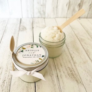 May include: Two small glass jars filled with white sugar scrub, tied with white ribbon and a personalised label. The label has a floral design and the text "Arielle and Jonathan September 28, 2029".