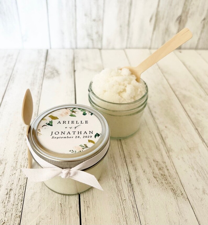 May include: Two small glass jars filled with white sugar scrub, tied with white ribbon and a personalised label. The label has a floral design and the text "Arielle and Jonathan September 28, 2029".