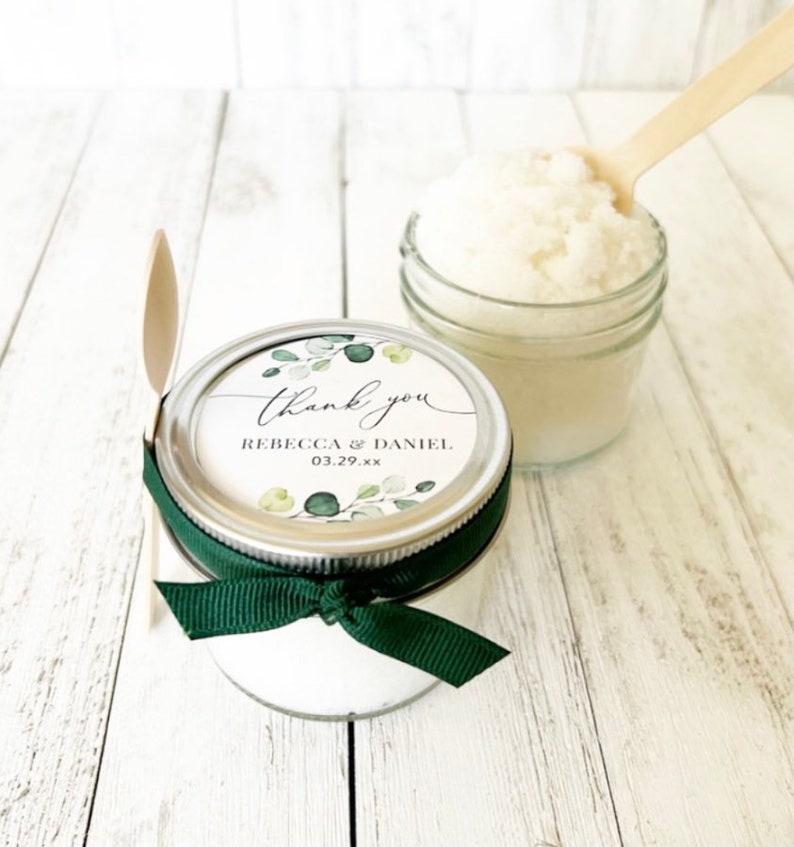 May include: A small glass jar filled with white sugar scrub, topped with a silver lid and a green and white sticker that says "thank you" with the names "Rebecca & Daniel" and the date "03.29.xx". A green ribbon is tied around the jar. The jar is sitting on a white wooden surface.