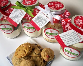 May include: Glass jars filled with cookie ingredients, labeled "Oatmeal Cookies", "Candy Chip Cookies", "Peanut Butter Cookies", and "Chocolate Chip Cookies". Each jar has a red lid with white snowflake pattern, a recipe tag, and a ribbon.