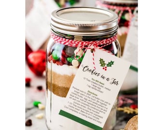 May include: A clear glass jar filled with layered ingredients for cookies, topped with a silver lid. The jar is decorated with red and white twine and a tag that reads "Cookies in a Jar". The jar contains chocolate chips and candies.