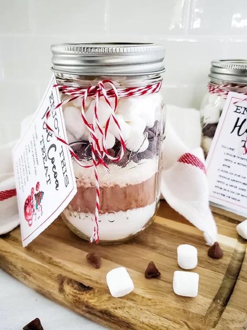 May include: A mason jar filled with layers of ingredients for hot cocoa, including marshmallows and chocolate chips. The jar is tied with red and white twine and has a tag that reads "Enjoy this Cocoa Treat". The jar sits on a wooden cutting board.