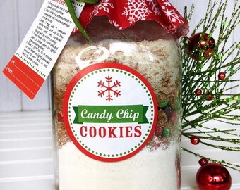 May include: A clear glass jar filled with cookie ingredients, labeled "Candy Chip Cookies." The jar is adorned with a red and white snowflake fabric lid, a green ribbon, and a tag with instructions. Festive holiday-themed gift.