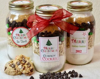 May include: Three glass jars filled with cookie ingredients, including oats, chocolate chips, and colorful candies. Each jar has a lid and a label that reads "Merry Christmas" and "Cowboy Cookies." One jar is tied with a red ribbon. A few cookies are in front.