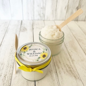 May include: Two glass jars filled with white sugar scrub, one with a yellow ribbon and a sunflower wedding label that reads "Jessica & William 10.25.XX", and the other with a wooden spoon.