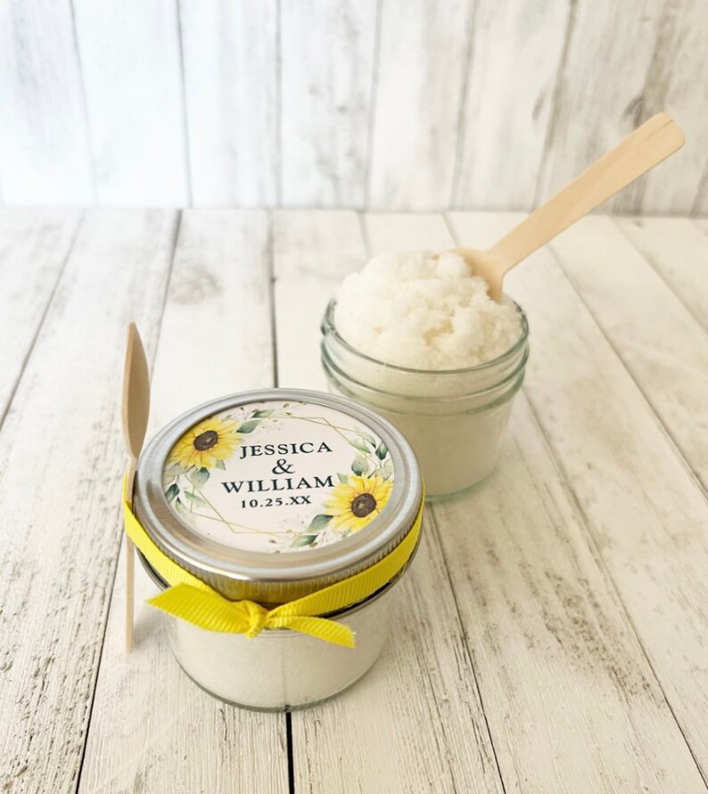 May include: Two glass jars filled with white sugar scrub, one with a yellow ribbon and a sunflower wedding label that reads "Jessica & William 10.25.XX", and the other with a wooden spoon.