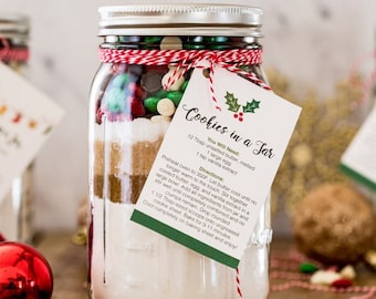 May include: A clear glass jar filled with layered ingredients for making cookies. The jar is tied with red and white twine and has a tag that reads "Cookies in a Jar." The jar contains chocolate chips, sprinkles, and other baking ingredients.