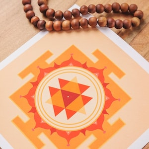 May include: A square print with a geometric design in orange and peach tones, a heart-shaped stone, and a wooden bead necklace are arranged on a wooden surface. The print features a central triangle design.