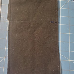 May include: A rectangular, black fabric item, possibly a case or pouch, is shown. The fabric appears to be a sturdy canvas or similar material. The item is folded over, with a visible seam running across the middle. The edges are neatly sewn.