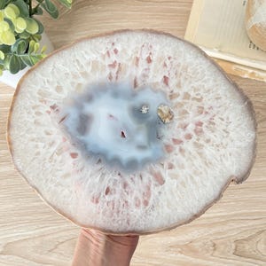 May include: A large, round agate slice with a light blue and white center, surrounded by a white and tan outer edge. The agate has a natural, organic appearance, with visible crystal formations. The slice is held by a hand.