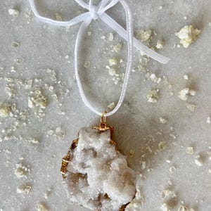 May include: A white crystal pendant with a gold rim, hanging from a white satin ribbon. The crystal has a rough, natural texture.