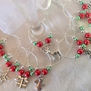 May include: Set of Christmas wine glass charms. Each charm features a silver-tone holiday-themed pendant, red and green beads, and a silver hoop. Charms include a star, angel, gift, snowman, snowflake, and reindeer.