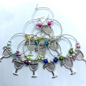 May include: A collection of silver-toned hoop wine glass charms, each featuring a textured cocktail glass charm and accented with colorful round beads in shades of pink, blue, green, yellow, and purple, arranged in a layered circular pattern.